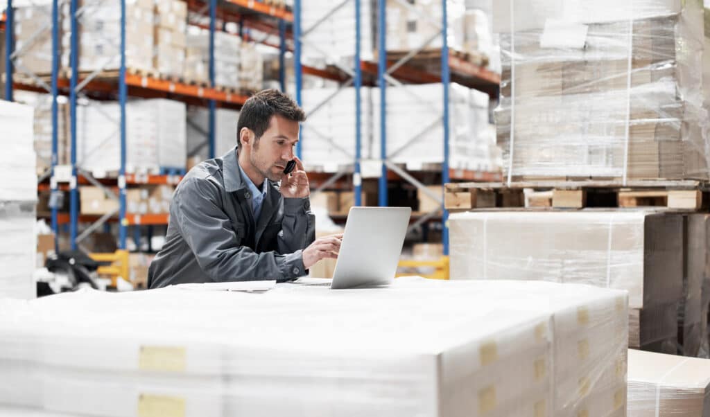 Man on phone looking at laptop computer in warehouse
